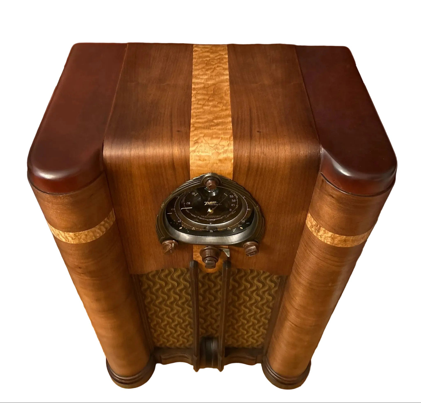 An antique wooden radio set with a polished mahogany finish, featuring a central wood grain inlay, a decorative metal dial, and a patterned front grille, positioned on a hardwood floor against a terracotta-colored wall.