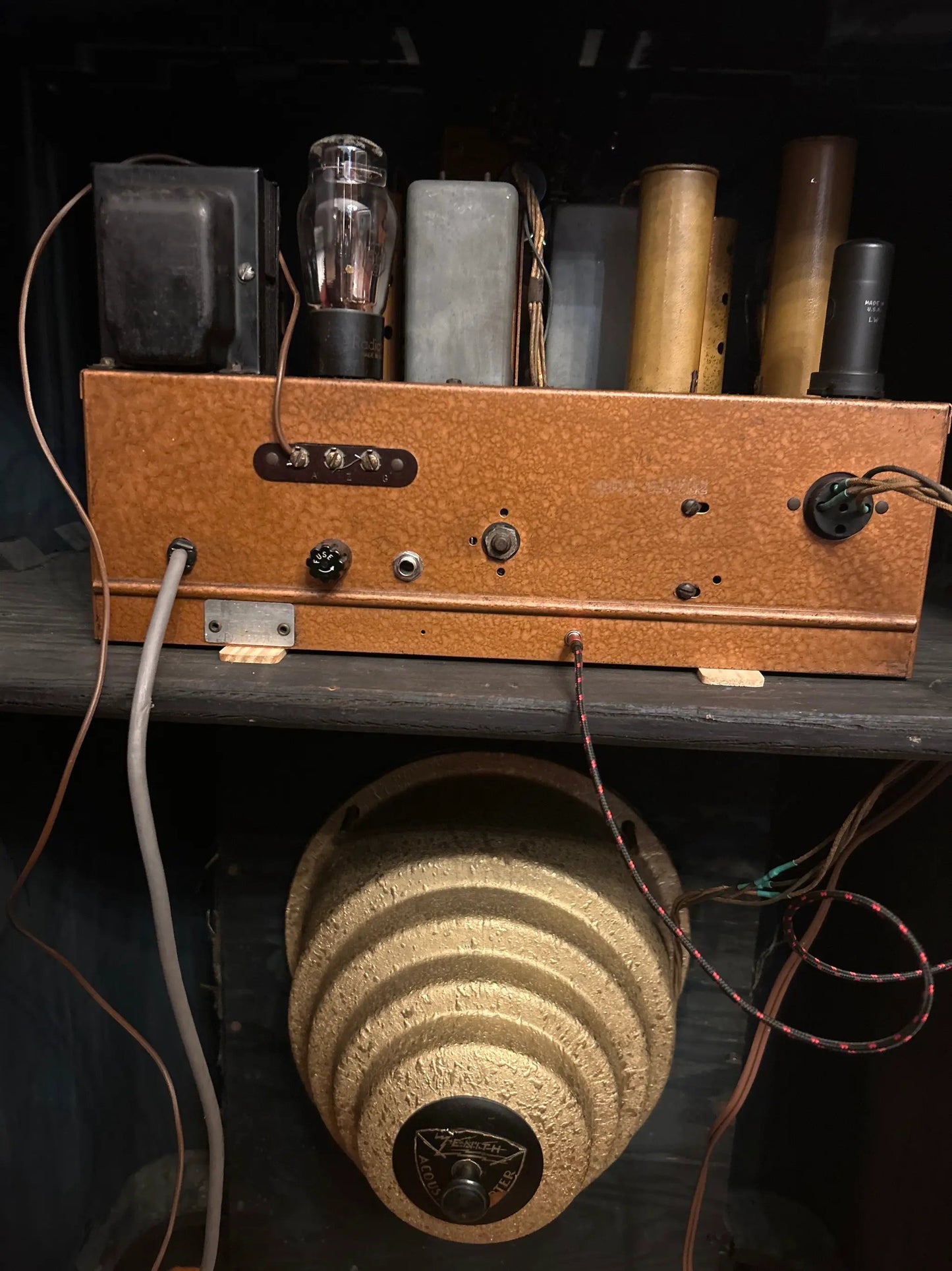 A vintage orange-brown wooden electronic amplifier or radio chassis with vacuum tubes and wires, sitting on a shelf above a large beige speaker cone.