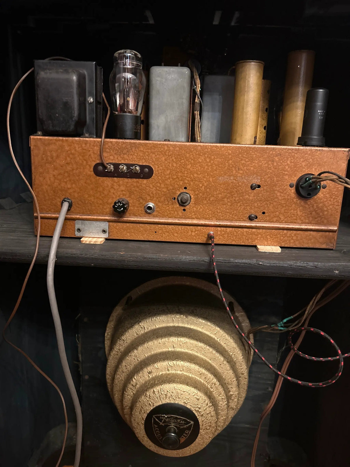 A vintage orange-brown wooden electronic amplifier or radio chassis with vacuum tubes and wires, sitting on a shelf above a large beige speaker cone.