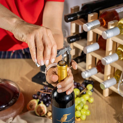 Person opening a bottle of wine with a corkscrew, surrounded by grapes and a wooden wine rack.